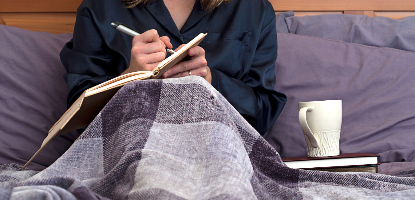 A woman sitting in her bed, covered by a blanket, embracing journaling by writing in her journal.