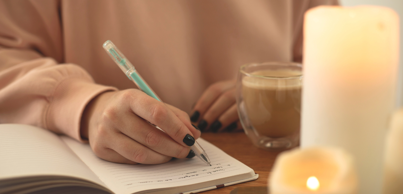 Closeup of a woman writing in a journal with candles burning and a cup of coffee nearby.