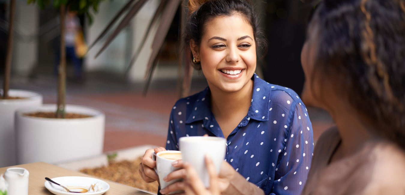 A woman smiling at another woman as they have coffee together.