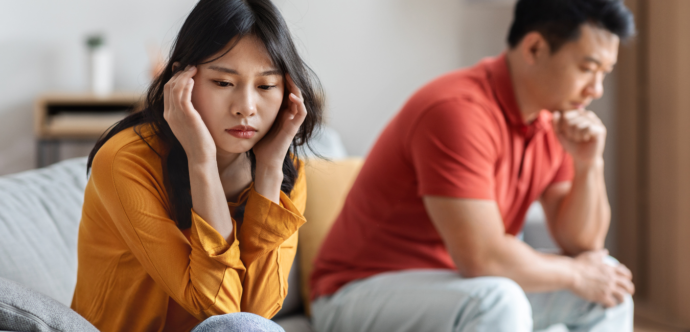 A woman sitting on a sofa with her ears plugged with her husband sitting behind her.