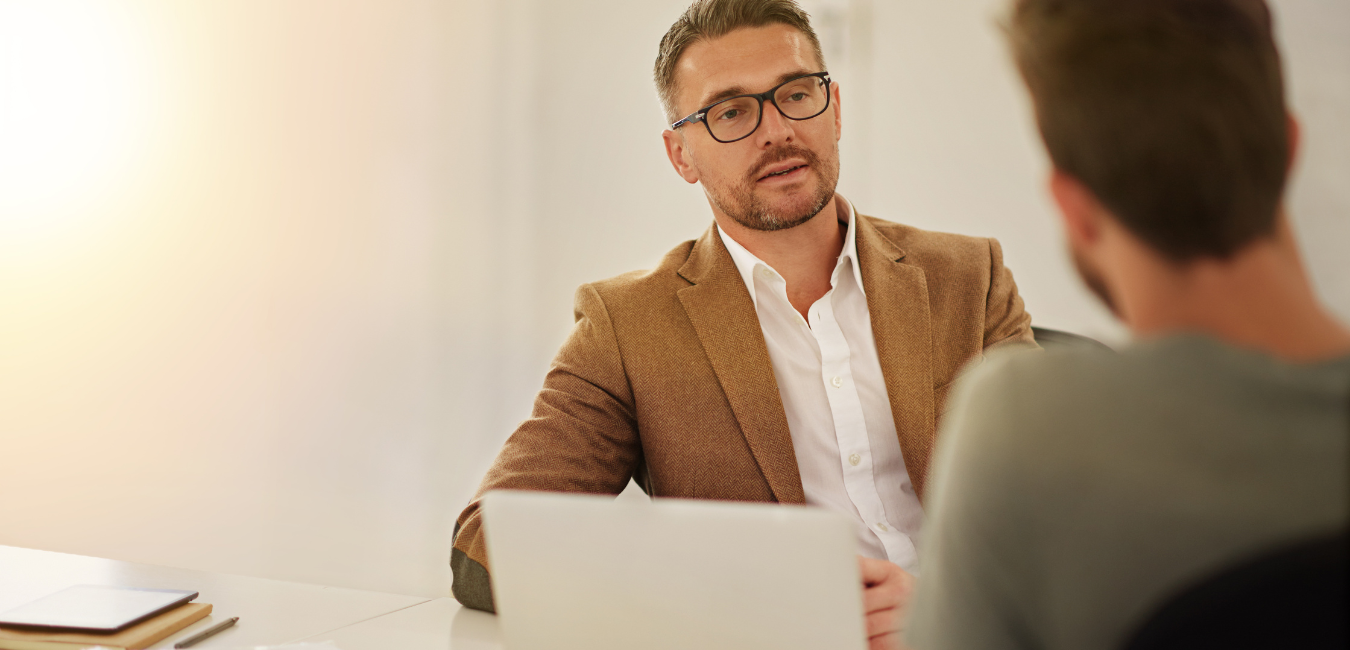 A man, sitting at a desk, talking to another man.