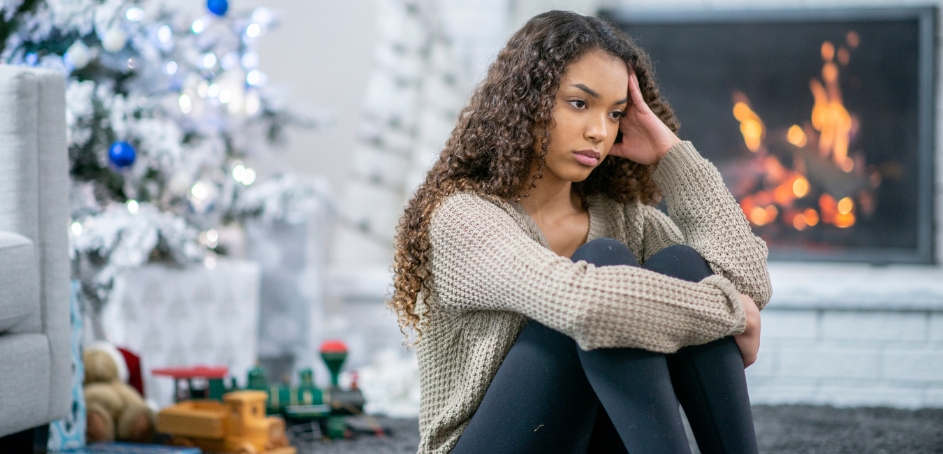 A young woman sitting in front of a Christmas tree, looking sad as she rests her hand on her hand.